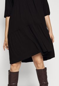 Svart lager-på-lager klänning med långa ärmar och asymmetrisk fåll, tillverkad av mjukt tyg. Kombinerad med knähöga bruna läderstövlar.