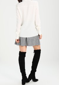 Žena v bílé blůze s dlouhým rukávem, šedé plisované sukni a černých kozách s vysokým podpatkem z semiše, stojící na jednobarevném pozadí.