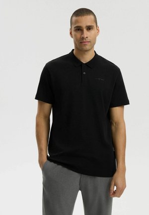 Mężczyzna o krótkich włosach, ubrany w czarną koszulkę polo i szare spodnie, stojący z jedną ręką w kieszeni na jednolitym tle.