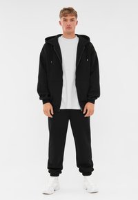 Zwarte zip-up hoodie met een losse pasvorm, gecombineerd met bijpassende zwarte joggers. Een witte t-shirt met ronde hals eronder. Gelaagd materiaal met ribbelmanchetten.