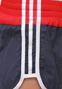 Pantaloni corti blu navy con una fascia elastica rossa in vita, caratterizzati da strisce verticali bianche lungo i lati e un design con orlo spaccato per maggiore mobilità.