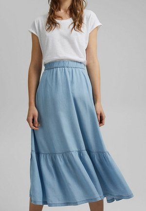 Femme portant un haut blanc à manches courtes et une jupe midi bleu clair, taille haute et à volants, sur un fond gris uni.