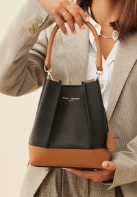 Black leather bucket bag with a tan base, structured shape, top handle, and gold branding. Features textured surface and metal hardware.