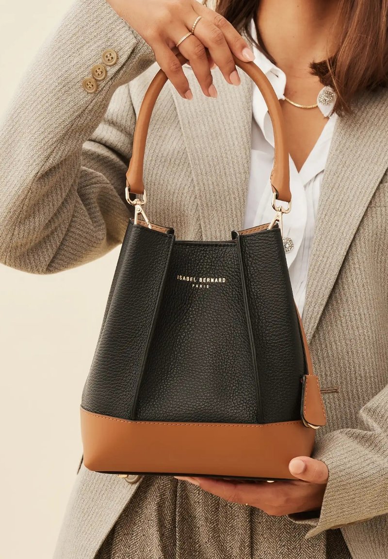 Black leather bucket bag with a tan base, structured shape, top handle, and gold branding. Features textured surface and metal hardware.