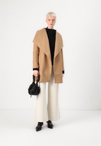Manteau camel avec col large, pull noir à côtes, pantalon large crème et petit sac à main noir à fleurs. Chaussures noires à bout pointu.