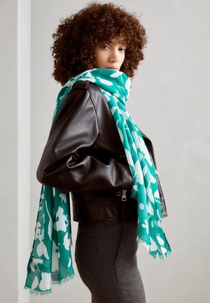 Femme aux cheveux bouclés portant une veste en cuir noire et une écharpe florale bleu sarcelle drapée sur les épaules, debout de profil devant un fond uni.