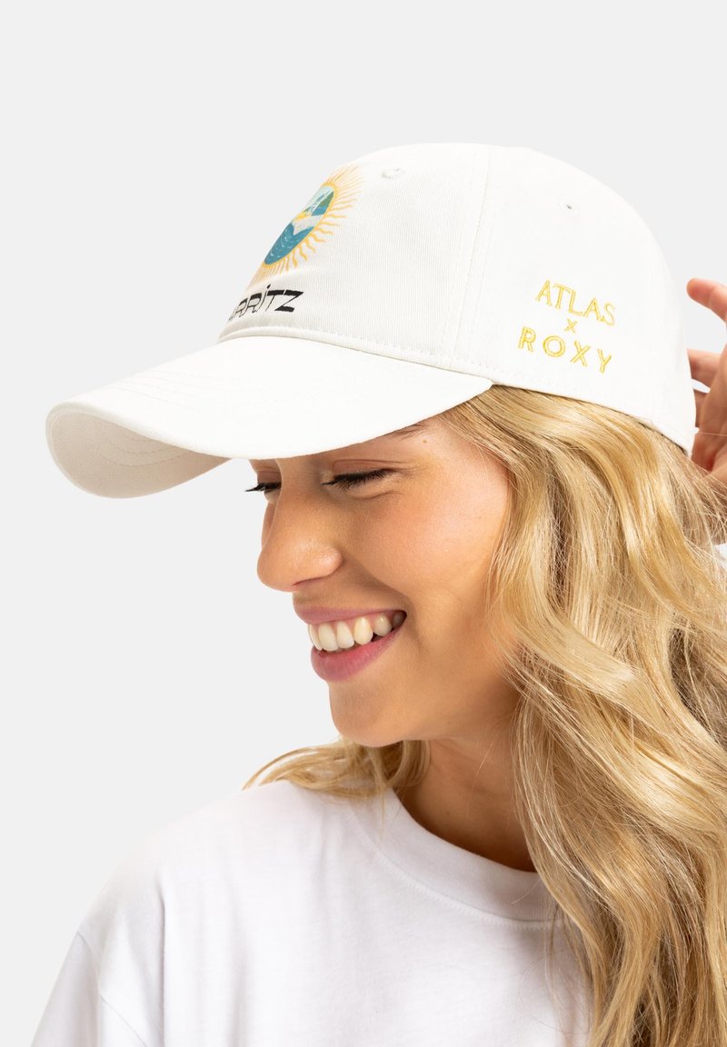 White cotton cap featuring colorful embroidered sun and mountain design at the front, with "ATLAS x ROXY" in gold on the side.