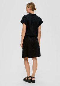 Robe en lin noire avec des manches courtes à revers, détail de poche arrière et taille élastique. Associée à des sandales noires à semelles épaisses.