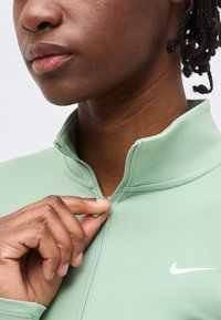 Άτομο με σκουρόχρωμο δέρμα και πλεγμένα μαλλιά που κουμπώνει ένα ανοιχτό πράσινο μπουφάν Nike με ανεβασμένο γιακά σε απλό φόντο.