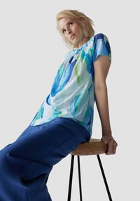 Blue and green patterned, short-sleeved top in a light, textured fabric, paired with a solid deep blue skirt, seated on a wooden stool.
