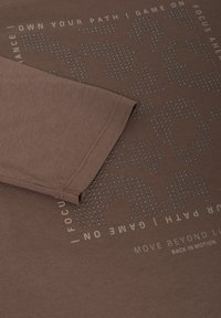 Brązowy bawełniany t-shirt z teksturowanym wzorem utworzonym przez małe wypukłe kropki. Nadrukowany tekst w kolorze kontrastującym wzdłuż jednej strony.