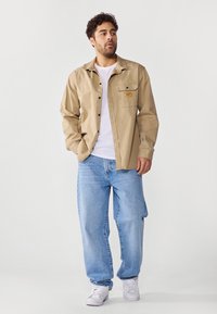 Camisa beige de botones con un bolsillo en el pecho y un parche de logo, usada sobre una camiseta blanca, combinada con unos vaqueros baggy de color azul claro y zapatillas blancas.