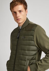 Veste bomber vert olive avec une partie avant rembourrée, texture lisse, fermeture éclair, col côtelé et poches latérales. Étiquette de la marque visible sur la poitrine.
