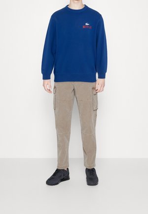 Modrá mikina s červeným logem "NETFLIX", béžové kapsáčové kalhoty a černé tenisky. Mikina má kulatý výstřih a žebrované manžety.