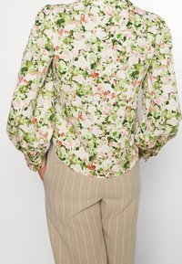 Květinová halenka z lehké látky s růžovými a zelenými vzory. Dlouhé rukávy s zaobleným lemem, kombinovaná s béžovými kalhotami s proužky.