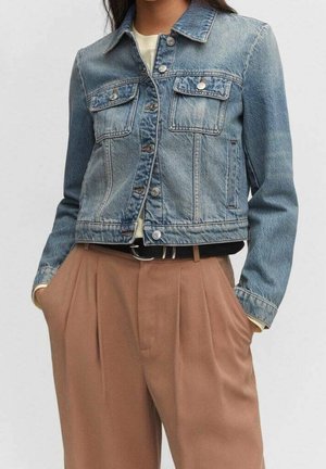 Person iført en blå denimjakke over en hvid skjorte, kombineret med høj talje, plisserede brune bukser og et sort bælte.