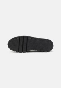 Zwarte rubberen schoenen zool met een getextureerd grippatroon en drie horizontale groeven voor flexibiliteit. Logo in reliëf aan de zijkant.
