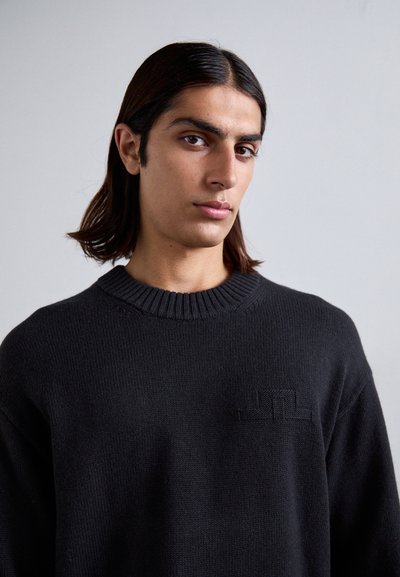 Black knitted sweater with a ribbed round neckline. Features a textured logo detail on the chest. Fabric appears soft and warm.