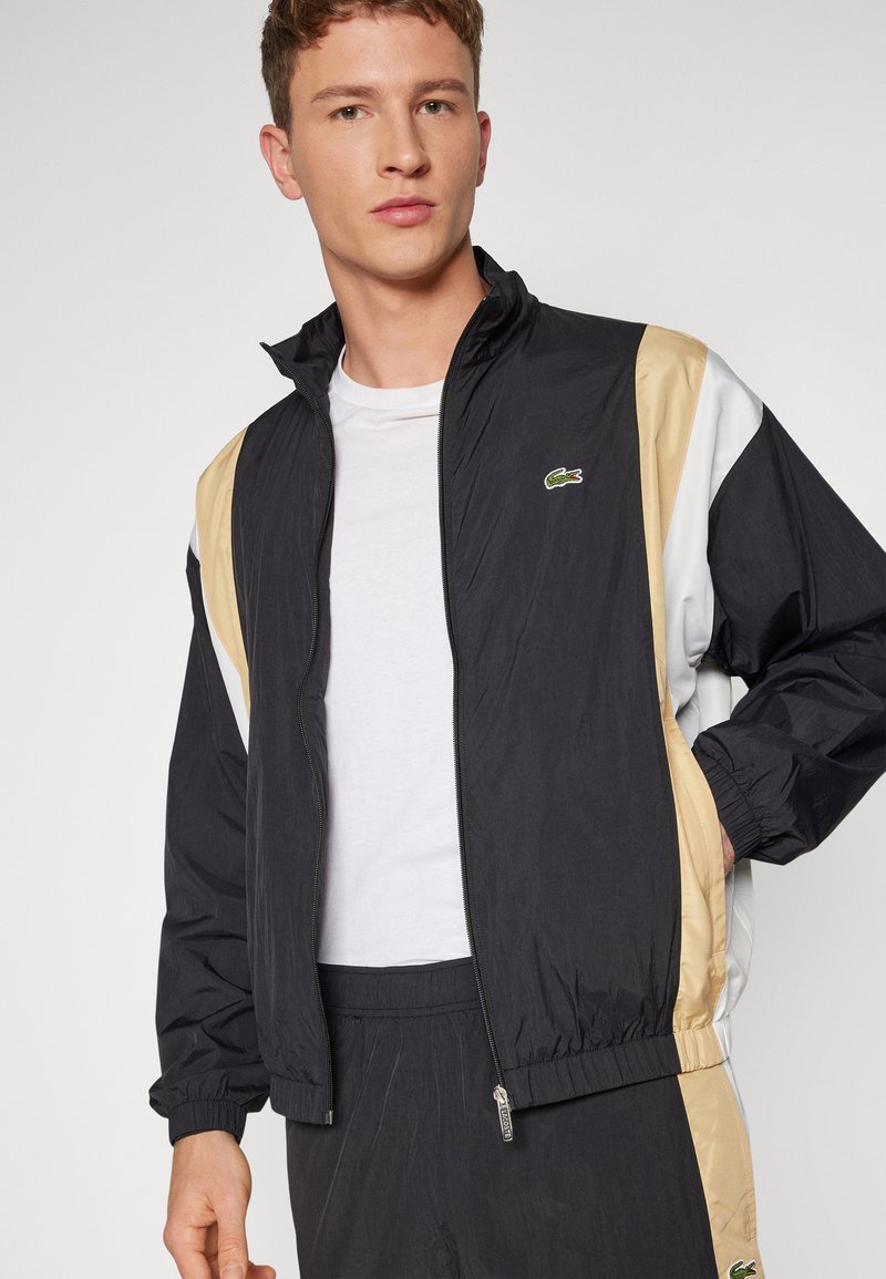 Veste noire, beige et blanche en tissu léger, dotée d'une fermeture éclair à l'avant, de poignets élastiques et d'un petit logo vert sur la poitrine.