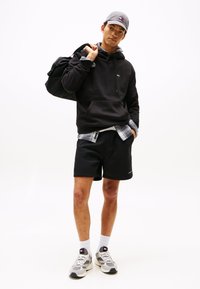 Camisola preta com cordões, boné cinza, camisa de padrão xadrez por baixo, calções pretos e ténis cinza com detalhes em branco. A levar uma mala preta.