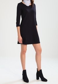 Vestido negro de manga larga en un tejido suave con escote redondo, combinado con botines negros. El conjunto incluye un accesorio de bufanda ligera.