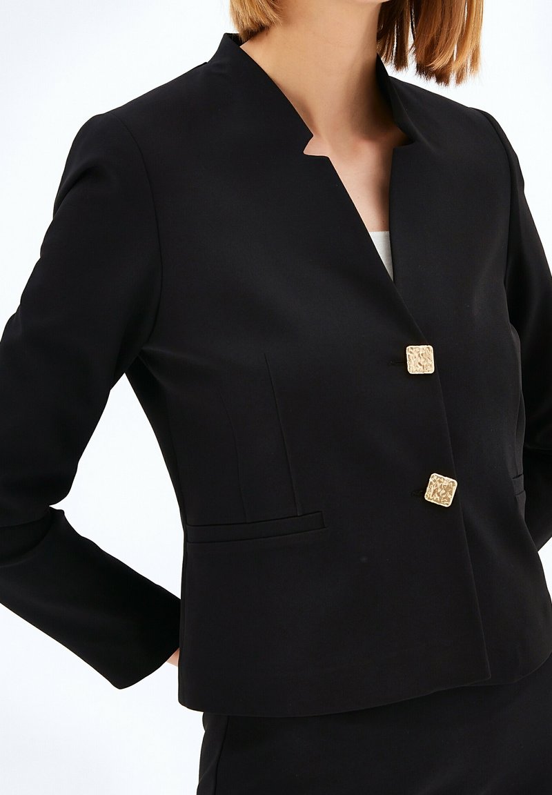 Donna che indossa un blazer nero su misura con due bottoni quadrati dorati texturizzati e un colletto a scalfo unico, abbinato a pantaloni neri.