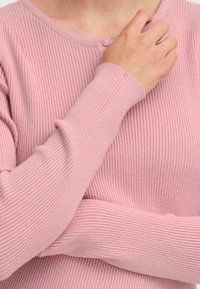 Ribbet lyserød sweater med lange ærmer og rund halsudskæring. Har en lille knapdetalje foran, der fremviser et blødt, struktureret stof.