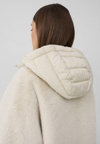Crèmekleurige, gestructureerde buitenkleding met een puffy, gewatteerde capuchon. Kenmerkt zich door een zachte, donzige stof, visueel warm en gezellig in ontwerp.