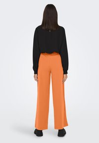 Zwarte lange mouw top met sleutelgat achter; oranje wijde broek; effen kleuren; gestructureerde stof; zwarte schoenen; eenvoudig ontwerp.