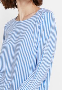 Kobieta ubrana w niebiesko-białą bluzkę w pionowe paski z długim rękawem i okrągłym dekoltem, pokazana od szyi do połowy torsu.