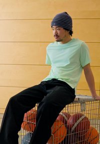 Lys grøn kortærmet skjorte, sorte løse bukser, sidder på en trådkurv fyldt med forskellige basketballer, stribet hue.
