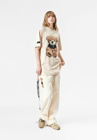 Camiseta oversized beige con un diseño de oso de peluche texturizado, combinada con pantalones color crema adornados con un parche de oso a juego. Calzado marrón.
