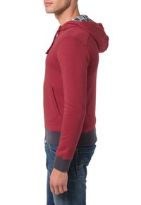 Felpa rossa con cappuccio realizzata in un misto cotone, presenta una tasca a marsupio, polsini e orlo a costine, con un rivestimento a scacchi visibile all'interno del cappuccio.