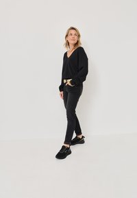 Černý svetr s výstřihem do V a dlouhými rukávy, černé slim džíny a černé tenisky se zlatými akcenty. Model stojí na neutrálním pozadí.