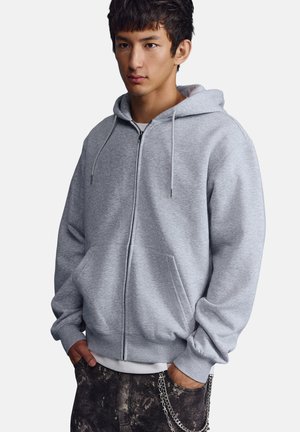 Mladý muž oblečený v svetlosivú mikinu na zips s kapucňou, ruky v vreckách a retiazka pripojená k tmavým vzorovaným nohaviciam, smerujúci vpred.