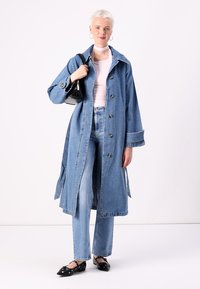 Trench coat in denim di colore blu chiaro, con chiusura a bottoni, fessure laterali e vestibilità oversize. Abbinato a jeans blu chiaro e ballerine nere.
