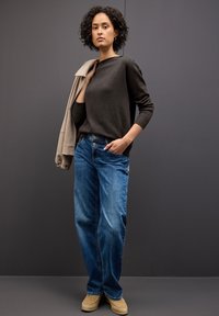Dark sweater with ribbed cuffs, blue straight-leg jeans, and beige slip-on shoes. Model holds a beige jacket against a gray background.
