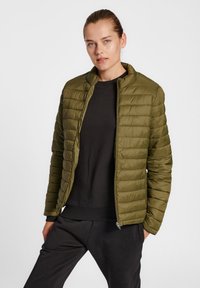Doudoune vert olive avec un design matelassé horizontal, un col haut et une fermeture éclair. Portée sur un sweat-shirt noir et un pantalon.