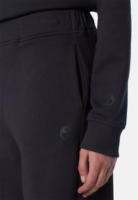 Pantalones de chándal negros con cintura ajustable mediante cordón, puños acanalados y dos bolsillos laterales. Presenta un sutil logotipo circular en el muslo y en la manga.