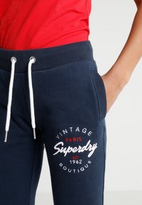 Mörkblå joggerbyxor i mjukt tyg med dragsko iMidjan; har logotyp med text i vitt och rött, ribbade mudd och sidofickor.