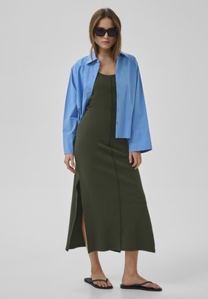 Nő sötétzöld ujjatlan maxiruhában, oldalhasítékkal, világoskék kigombolatlan ingben, fekete napszemüvegben és fekete papucsban szürke háttér előtt áll.