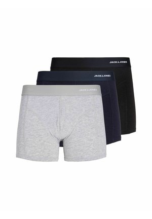 Jeune homme debout, torse nu, portant un boxer gris clair avec une ceinture grise sur un fond uni.