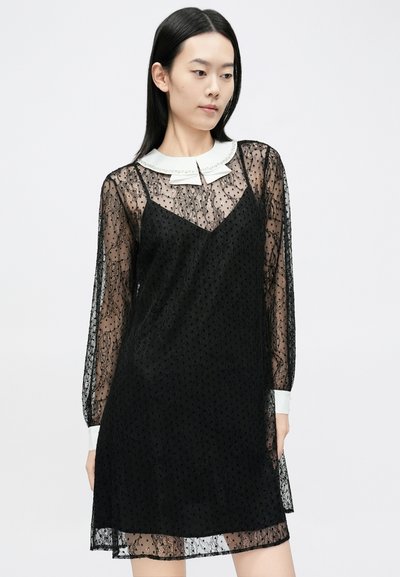 Svart blondekjole med et gjennomsiktig lag, lange ermer og en hvit krage med dekorative detaljer. Litt A-formet silhuett.