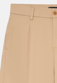 Beiga skräddarsydda byxor med en slät yta, en framficka och en enda brun knappstängning i midjan.