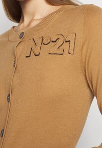 Hnědý pletený cardigan s kulatým výstřihem, vybavený zapínáním na knoflíky a černým vyšitým nápisem "N°21" na levé straně.