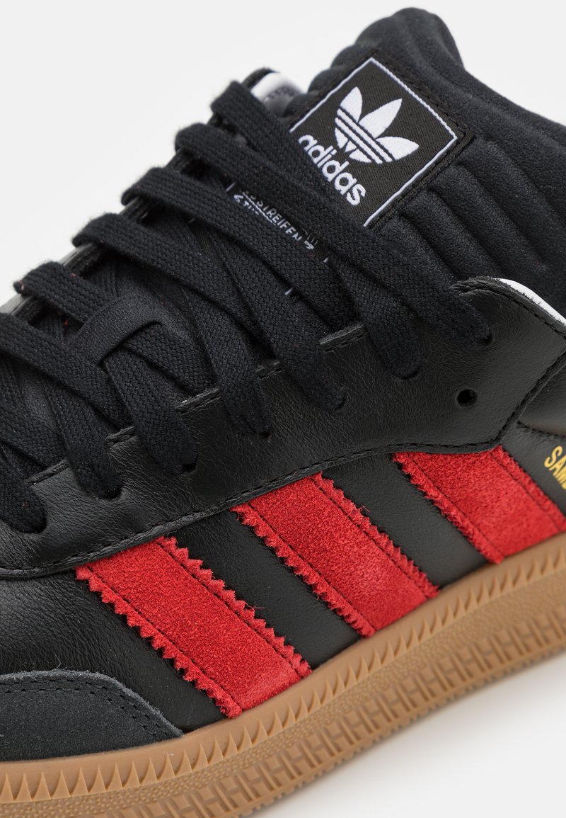 Czarne skórzane sneakersy z czerwonym zamszowym trzema paskami, teksturowanym czarnym językiem z logo i gumową podeszwą. Teksturowane akcenty po bokach.