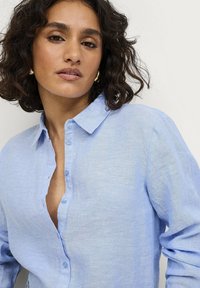 Camisa de lino azul claro con botones y cuello, que presenta un frente parcialmente desabrochado y una tela texturizada. Corte suave y relajado.