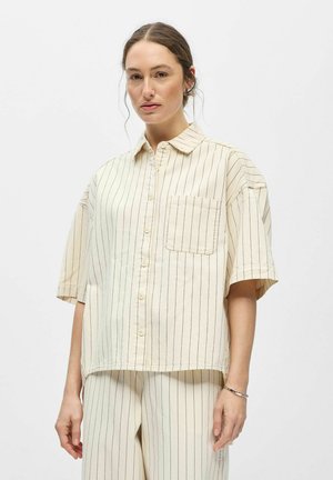 Femme portant une chemise crème à manches courtes boutonnée et un pantalon assorti avec de fines rayures verticales noires sur fond blanc.