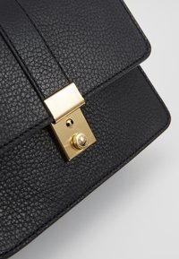 Borsa a tracolla nera in pelle texturizzata con una chiusura a scatto dorata e dettagli in oro. Presenta una chiusura a patta e cuciture visibili lungo i bordi.