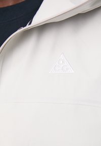 Ljust beige jacka med en slät textur, som har en vit triangulär logotyp på bröstet och en dragkedjestängning.